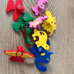 Puede incluir: Un conjunto de coloridos pomos de madera con forma de dinosaurio en rojo, rosa, verde, amarillo y azul. Cada pomo tiene un pequeño tornillo redondo de color plateado. Los pomos están dispuestos sobre una superficie de madera marrón claro.