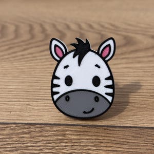 Peut inclure: Épingle en forme de tête de zèbre blanc et noir, avec un museau gris et des rayures noires. L'épingle a un contour noir et des accents roses sur les oreilles. Le zèbre a une expression souriante. L'épingle est probablement en plastique.