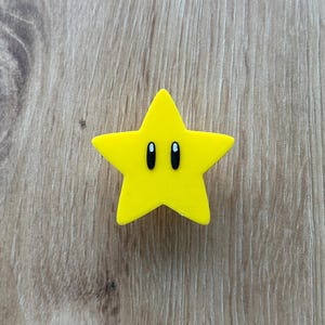 Op de afbeelding: Een heldergele stervormige bedel met zwarte ovale ogen. De bedel is effen van kleur en heeft een glad oppervlak. De ster is een populair videogame-personage.