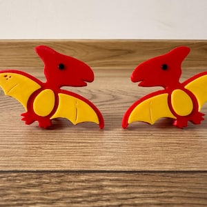 Può includere: Due ornamenti a forma di pterodattilo rossi e gialli. Ognuno ha una testa e un corpo rossi con ali gialle. Le ali hanno una forma curva. Gli ornamenti sono su una superficie di legno.