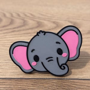 Puede incluir: Un pin gris con forma de elefante con contornos negros y orejas internas rosas. El elefante tiene ojos negros, mejillas rosadas y una trompa enroscada. El pin está sobre un fondo de madera.