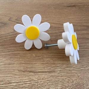 Può includere: Due pomelli per armadi a forma di margherita bianca con centri gialli. I pomelli sono fissati a una vite argentata.