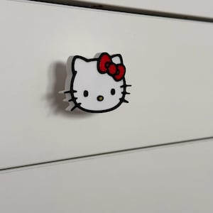 Op de afbeelding: Witte plastic ladetrekker met een zwarte omtrek van Hello Kitty's gezicht en een rode strik.