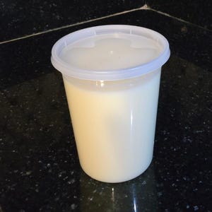 May include: A clear plastic container with a white lid, filled with a creamy, off-white liquid. The container is cylindrical and appears to be holding a generous amount of the substance. The lid is securely in place.