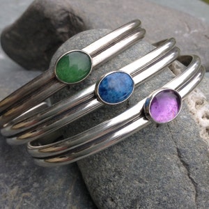 May include: Three silver bangle bracelets with oval-shaped gemstones. The bracelets are stacked on top of each other. The gemstones are green, blue, and purple.