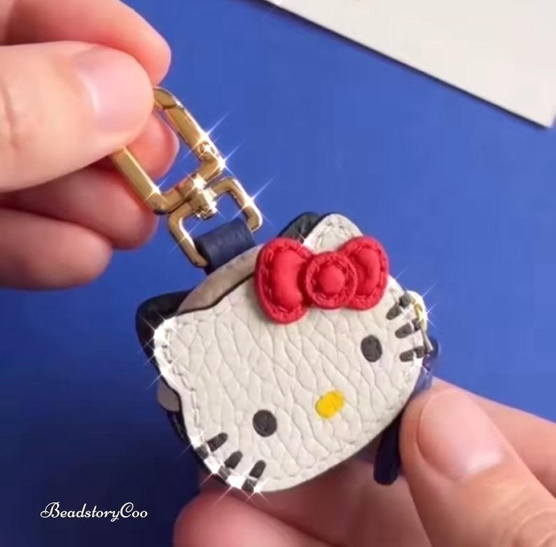 May include: A white, black, and red Hello Kitty keychain with a gold-colored clasp. The keychain features a red bow and black eyes. The text "BeadstoryCoo" is visible in the bottom left corner.