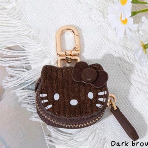 May include: Dark brown corduroy coin purse shaped like a cartoon cat face with a bow. The purse has a gold-colored zipper and a matching key ring clasp. The text "Dark brown" is at the bottom of the image.