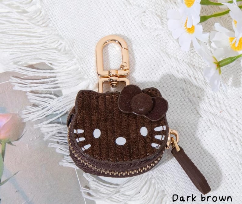May include: Dark brown corduroy coin purse shaped like a cartoon cat face with a bow. The purse has a gold-colored zipper and a matching key ring clasp. The text "Dark brown" is at the bottom of the image.