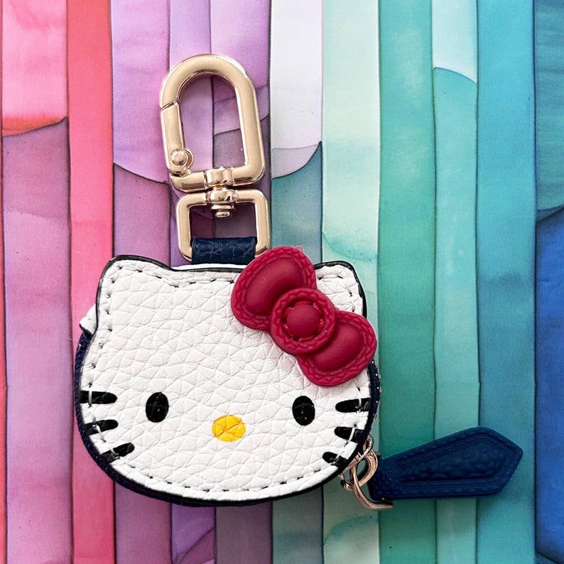 May include: White leatherette keychain featuring a Hello Kitty design with a red bow and black eyes. The keychain has a gold-tone clasp and a navy blue accent. The background is a colorful gradient of pink, purple, blue, and green.
