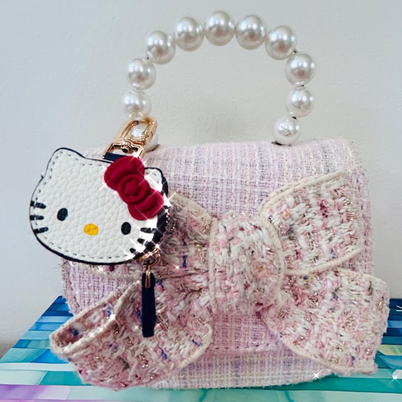 May include: A light pink tweed handbag with a large bow and a pearl handle. The bag features a Hello Kitty charm with a red bow. The bag is a small, stylish accessory.