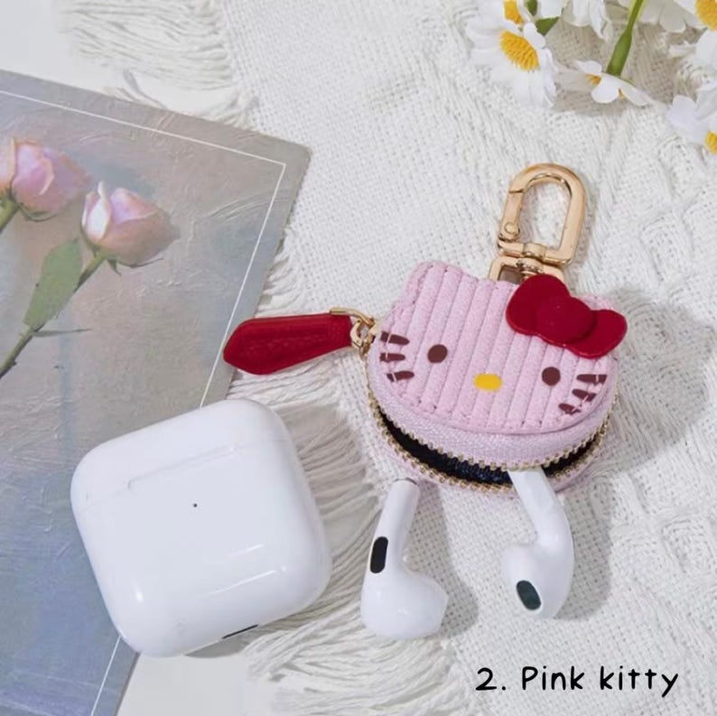 May include: A pink corduroy case shaped like a cartoon cat head with a red bow, black eyes, and yellow nose. The case is attached to a gold-colored clasp and holds white earbuds. A white charging case is also present.