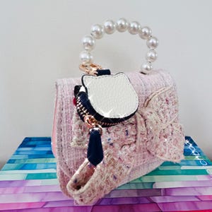 May include: A pink tweed handbag with a pearl handle and a decorative bow. The bag features a white cat-shaped charm with a black zipper and a navy blue tassel. The bag is sitting on a stack of colorful paper.
