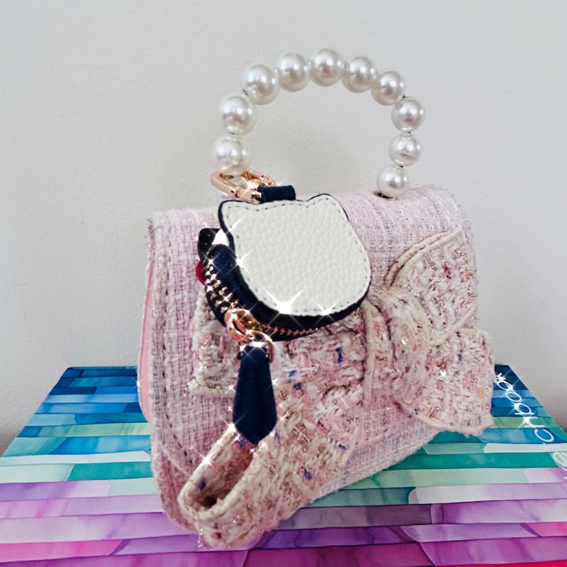 May include: A pink tweed handbag with a pearl handle and a decorative bow. The bag features a white cat-shaped charm with a black zipper and a navy blue tassel. The bag is sitting on a stack of colorful paper.