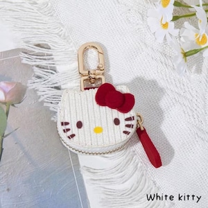 May include: A white, corduroy-textured coin purse in the shape of a cat's face, featuring a red bow, brown whiskers, and a yellow nose. It has a gold-colored clasp and a red zipper pull. The text "White kitty" is at the bottom.
