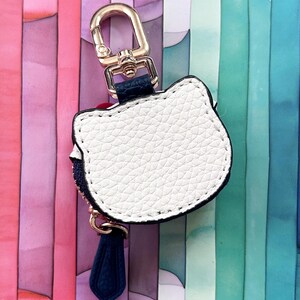 May include: White cat-shaped keychain with a gold-tone clasp and navy blue tassel. The keychain has a textured surface and a black border. The background features vertical stripes of pink, blue, and green.