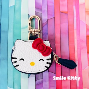 May include: A white and black Hello Kitty keychain with a red bow and gold-colored hardware. The keychain has a navy blue tassel and the text "Smile Kitty" in pink.