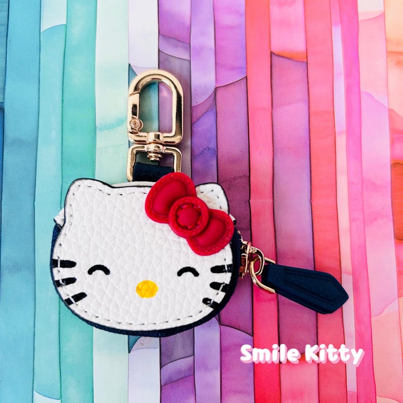 May include: A white and black Hello Kitty keychain with a red bow and gold-colored hardware. The keychain has a navy blue tassel and the text "Smile Kitty" in pink.