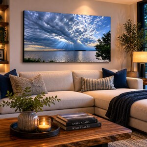 May include: A large framed canvas print of a dramatic sky over a body of water. The sky is filled with dark, swirling clouds, with beams of sunlight breaking through. A tree is visible on the right side of the image. The print is displayed above a beige sectional sofa.