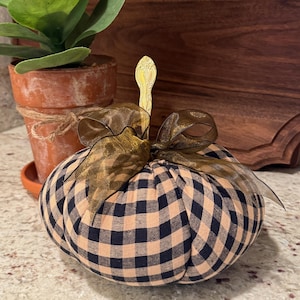 Op de afbeelding: Een stoffen pompoen met een beige en marineblauw ruitpatroon. Het heeft een goudkleurige steel en een bruine strik. De pompoen is een decoratief item, waarschijnlijk voor herfstdecoratie.