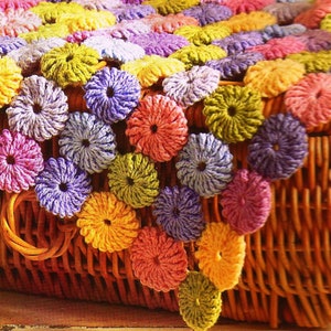 Pode incluir: Uma manta de crochê feita de círculos coloridos em tons de roxo, rosa, amarelo e verde. A manta está drapeada sobre uma cesta de vime.