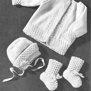 May include: A white knitted baby cardigan, hat, and booties set. The cardigan has a button closure and a textured stitch pattern. The hat has a bow and the booties have a ribbon tie.
