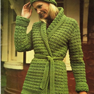Könnte beinhalten: Eine grüne gehäkelte Jacke mit passendem Hut. Die Jacke hat einen Schalkragen und einen Bindegürtel. Der Hut ist ein einfacher Beanie-Stil.