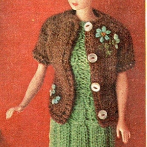May include: A doll wearing a green knitted dress and a brown knitted bolero jacket with white buttons and floral embellishments. The doll has blonde hair and is posed against a red background. The outfit is a vintage style.