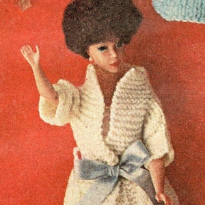 May include: A doll wearing a white crocheted dress with a blue satin bow. The doll has dark brown hair and is wearing earrings.