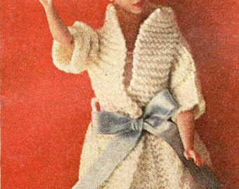 May include: A vintage doll in a cream-colored crocheted dress with a matching shawl and a light blue bow at the waist. The doll has a dark brown fur hat and is set against a red background.