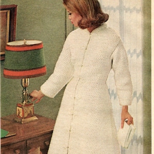 May include: A woman in a white knitted robe stands in a room with a lamp and a plant. She is holding a white book in her right hand.