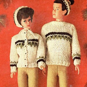 Op de afbeelding: Twee poppen in bijpassende gebreide outfits. De outfits omvatten crèmekleurige truien en jasjes met zwarte en groene accenten, gecombineerd met een beige broek. Eén pop draagt een bijpassende hoed. De achtergrond is effen rood.