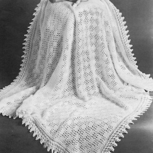 Puede incluir: Una manta de bebé de ganchillo blanco con un delicado borde de encaje. La manta está drapeada sobre una muñeca.