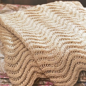May include: A beige knitted throw blanket with a wave pattern. The blanket is folded over, revealing the intricate stitchwork and texture. The text "This soothing wave pattern throw is a great way to wrap up your day!" is visible.