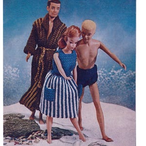 Op de afbeelding: Vintage afbeelding van drie poppen op een strandscène. Eén pop draagt een bruine badjas, een andere een blauw-wit gestreepte jurk met een blauwe top en zak, en de derde pop draagt een blauwe zwembroek. De achtergrond is een lichtblauwe lucht.