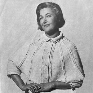 May include: A black and white image of a woman wearing a knitted cape. The cape is light-colored, buttoned down the front, and has a collar. The cape has a textured design with vertical lines and small holes. The woman is smiling.