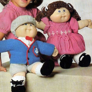 May include: Two Cabbage Patch Kids dolls wearing knitted outfits. The boy doll is wearing a blue sweater with a red trim and a white sailor collar, grey trousers, and a grey hat. The girl doll is wearing a pink dress with white embroidery and a white bow. Both dolls are wearing black shoes.
