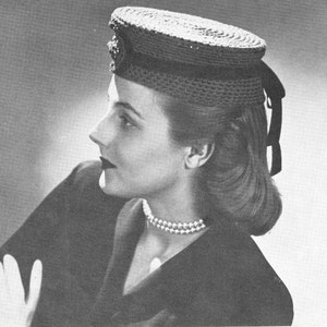 May include: A black and white photo of a woman wearing a crocheted hat with a black ribbon. The hat is a round, flat shape with a brim. The woman is wearing a dark jacket and a pearl necklace.