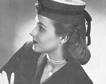 May include: A black and white photo of a woman wearing a crocheted hat with a black ribbon. The hat is a round, flat shape with a brim. The woman is wearing a dark jacket and a pearl necklace.