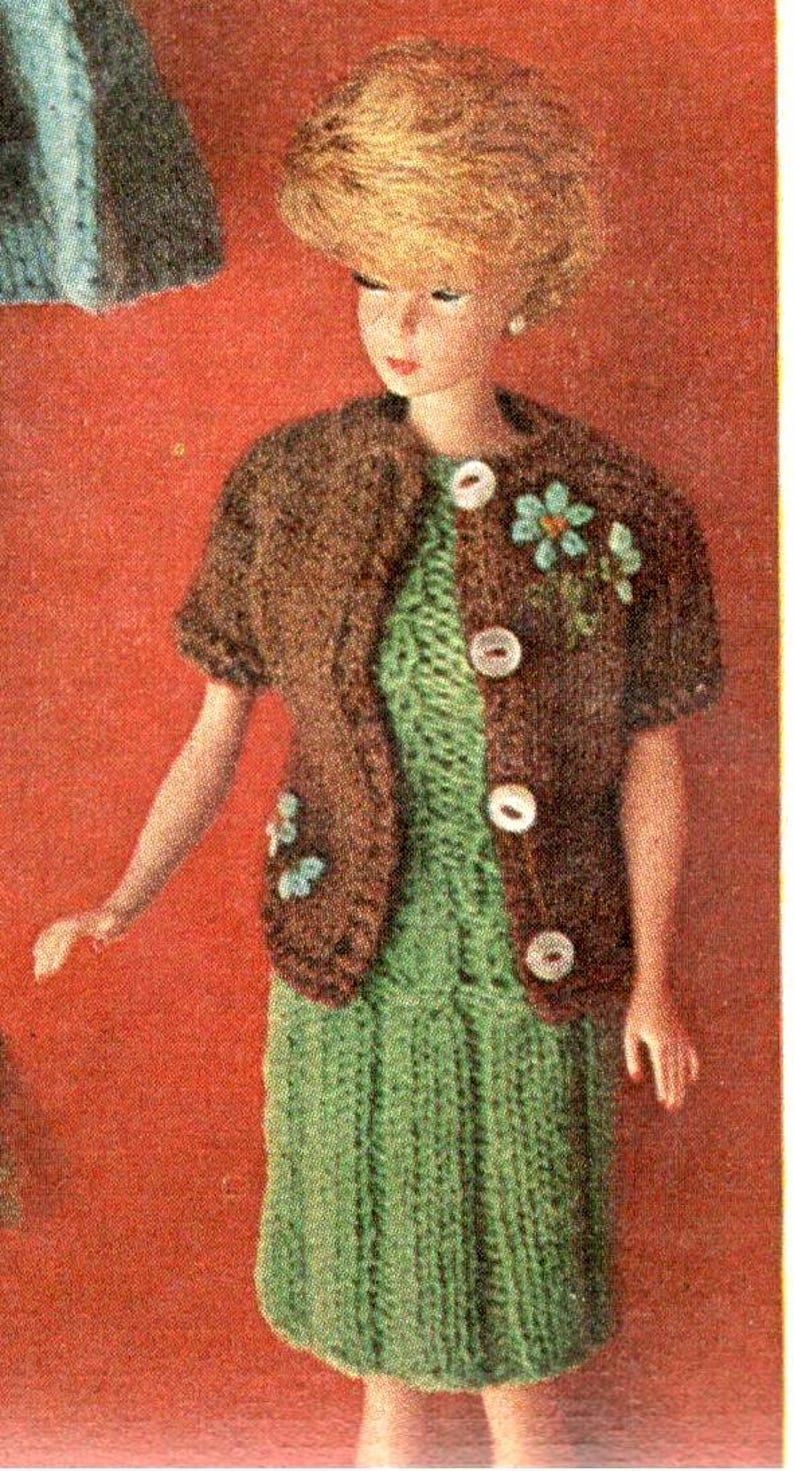 Puede incluir: Mu&ntilde;eca vestida con un atuendo hecho a mano. El conjunto incluye un vestido de punto verde y una chaqueta bolero marr&oacute;n con botones blancos y adornos florales. La mu&ntilde;eca tiene el pelo rubio, sobre un fondo rojo.