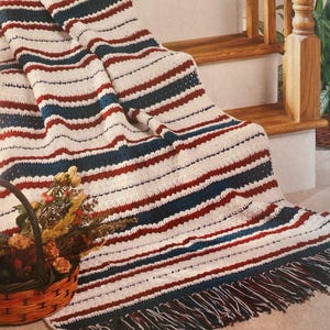 May include: A crocheted blanket with horizontal stripes in white, navy blue, and burgundy. The blanket has a fringed edge and is draped over a staircase. A basket of flowers sits in the foreground.