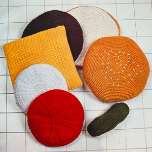 May include: A collection of handmade crochet food items, including a red tomato, a green pickle, and a round orange burger bun with white seed details. Other items include a yellow square pillow, and white, brown, and tan round cushions.