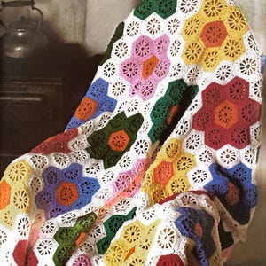 May include: A colorful crocheted blanket draped over a chair. The blanket features a repeating pattern of hexagonal shapes in various colors, including red, yellow, green, blue, and pink, with white floral accents. The blanket is made of yarn.