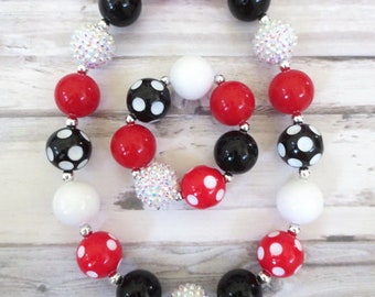 May include: A necklace with large, round beads in red with white polka dots, black, white, and sparkling silver. The beads are strung together to form a circular shape. The necklace is on a light-colored wooden surface.