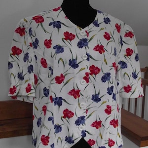 May include: A white button-up blouse with a floral print featuring red, blue, and green flowers. The blouse has a V-neck and short sleeves.