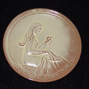 Peut inclure: Une assiette en céramique avec une image en relief d'une femme cousant. L'assiette est beige avec une bordure brune et les mots "Dorcas the Seamstress" sont écrits autour du bord inférieur.