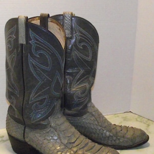 May include: A pair of cowboy boots with a gray snakeskin pattern on the foot and a black leather upper. The boots have decorative stitching in blue and gray, with pull straps at the top. The heels are dark brown.