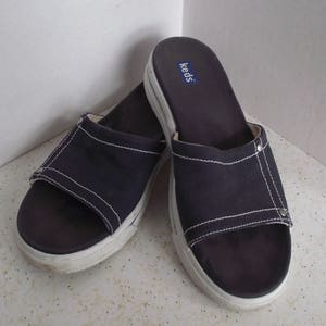 Op de afbeelding: Een paar donkerblauwe Keds slip-on sandalen met witte stiksels. De sandalen hebben een canvas bovenwerk en een witte rubberen zool.