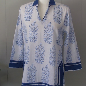 Op de afbeelding: Een witte tuniek met een blauwe bloemenprint en blauwe bies. De tuniek heeft een V-hals en driekwart mouwen.