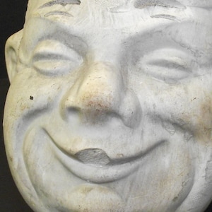 Puede incluir: Primer plano de una escultura de yeso blanco de un rostro con una amplia sonrisa de dientes. La escultura tiene una superficie texturizada y parece envejecida.