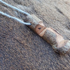 May include: A wooden pendant necklace with a natural bark finish. The pendant is shaped like a small, irregular cylinder with a carved design on one side. The necklace is made of a thin, light blue cord.